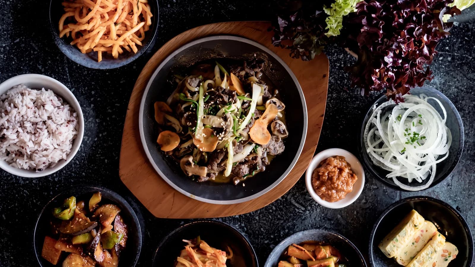 Bulgogi im Topf, von oben fotografiert, umgeben von Reis, Kimchi und weiteren Banchan
